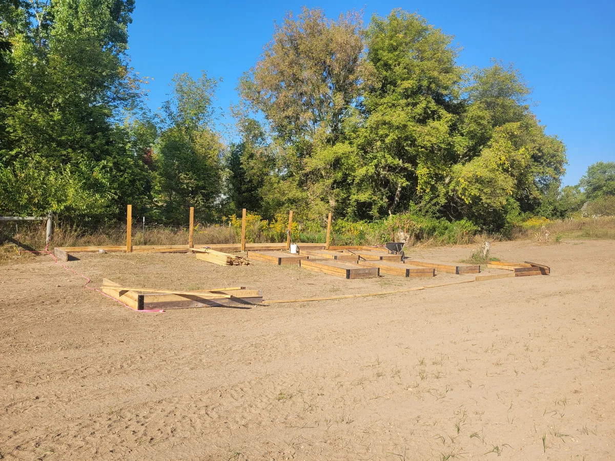 Wooden garden-bed frames and upright fence posts laid out on open ground beside trees.
